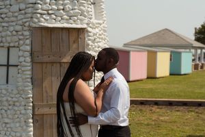A romantic couple shares an intimate moment beside a stone wall with colorful beach huts in the background.