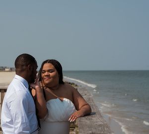 Romantic couple embraces while standing on a wooden deck overlooking a serene beach at sunset.