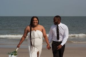 A couple in formal attire walks hand in hand along the beach with ocean waves behind them.