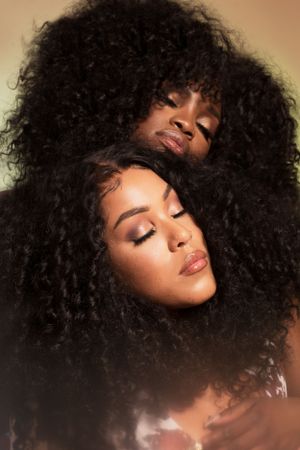 Beautiful portrait of two models with voluminous natural curly afro hairstyles posing with closed eyes in warm lighting.
