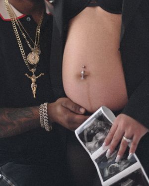 Person wearing a crucifix necklace shows an ultrasound photo while displaying a bare midriff with a navel piercing.