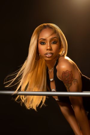 Portrait photo featuring long blonde hair, bold makeup, tattoo art and gold jewelry against dark dramatic lighting.