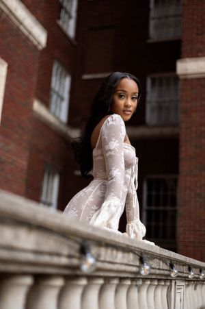 Elegant model in white lace dress poses on stone balcony against brick building background.