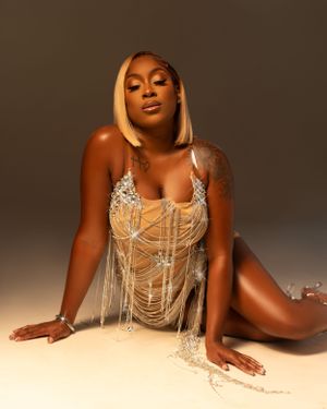 Model poses on floor wearing shimmering beige bodysuit with crystal embellishments and draping chains against tan backdrop.