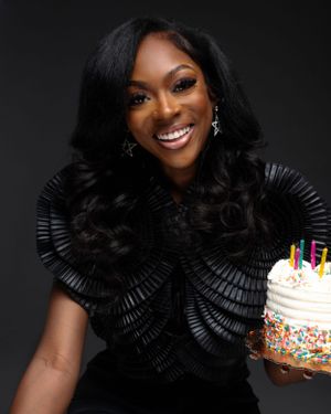A joyful portrait against a dark background showing someone in a black ruffled top holding a colorful birthday cake.