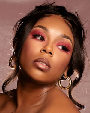 Close-up beauty photo featuring dramatic pink eyeshadow, gold hoop earrings and glossy nude lips in warm lighting.