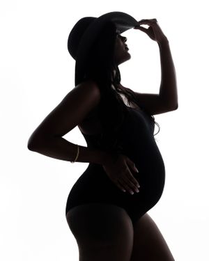 Silhouetted figure in a maternity pose wearing a hat touches the brim while standing against a white background.