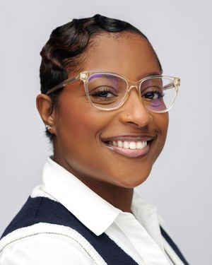 Professional portrait showing clear-framed glasses, a white collared shirt and navy blazer against a light background.