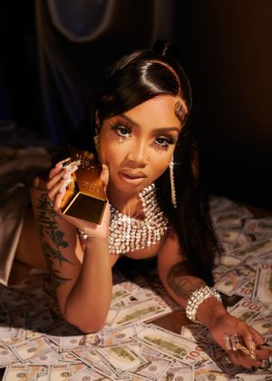 Queen Leora wearing diamond jewelry poses on bed with cash and gold bar in glamorous photoshoot.