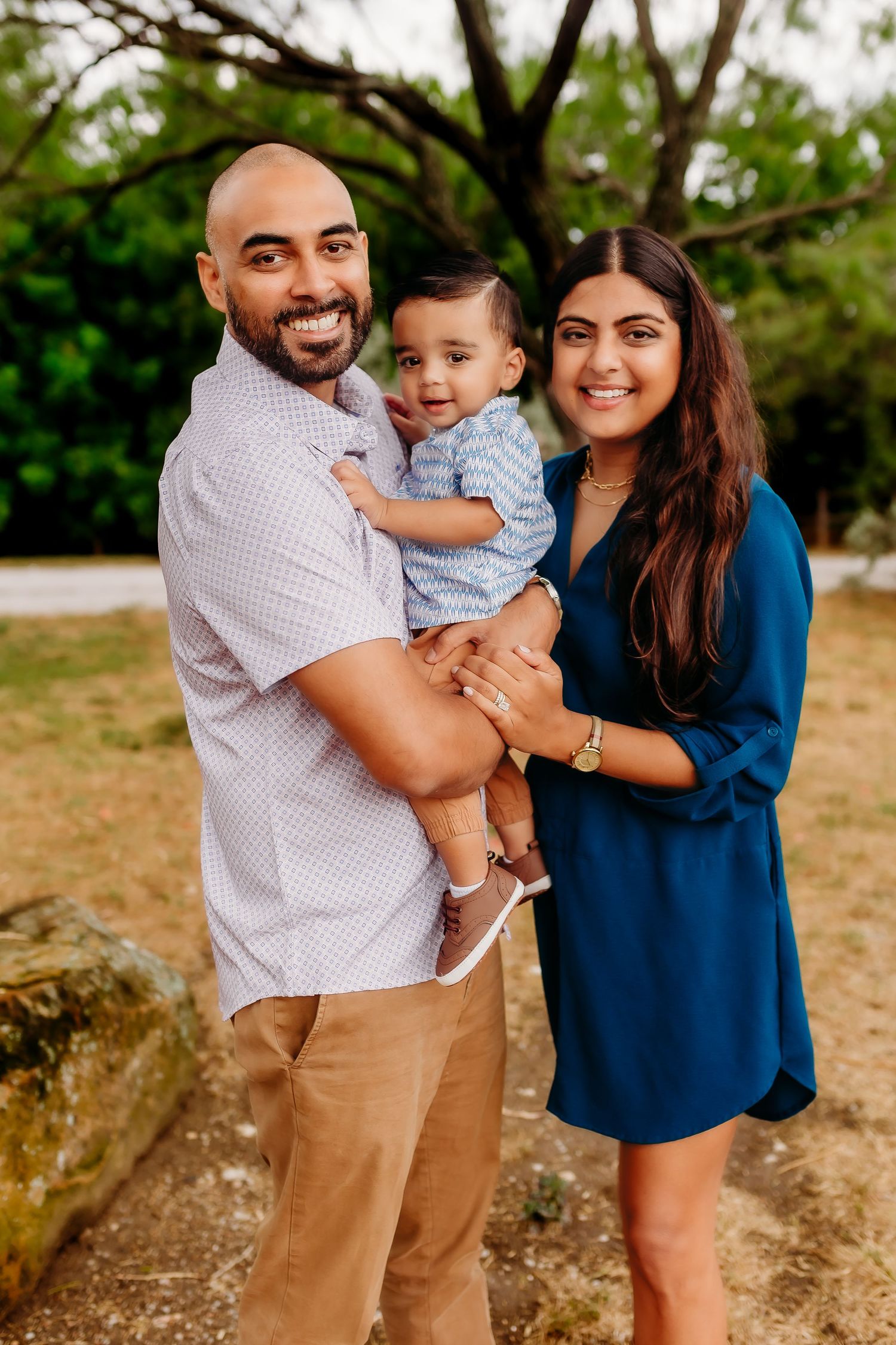 A family of three poses outdoors against a natural backdrop in coordinated outfits.