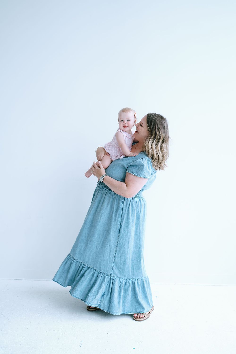 Mother in blue maxi dress holding and kissing her baby girl during professional studio photo session.
