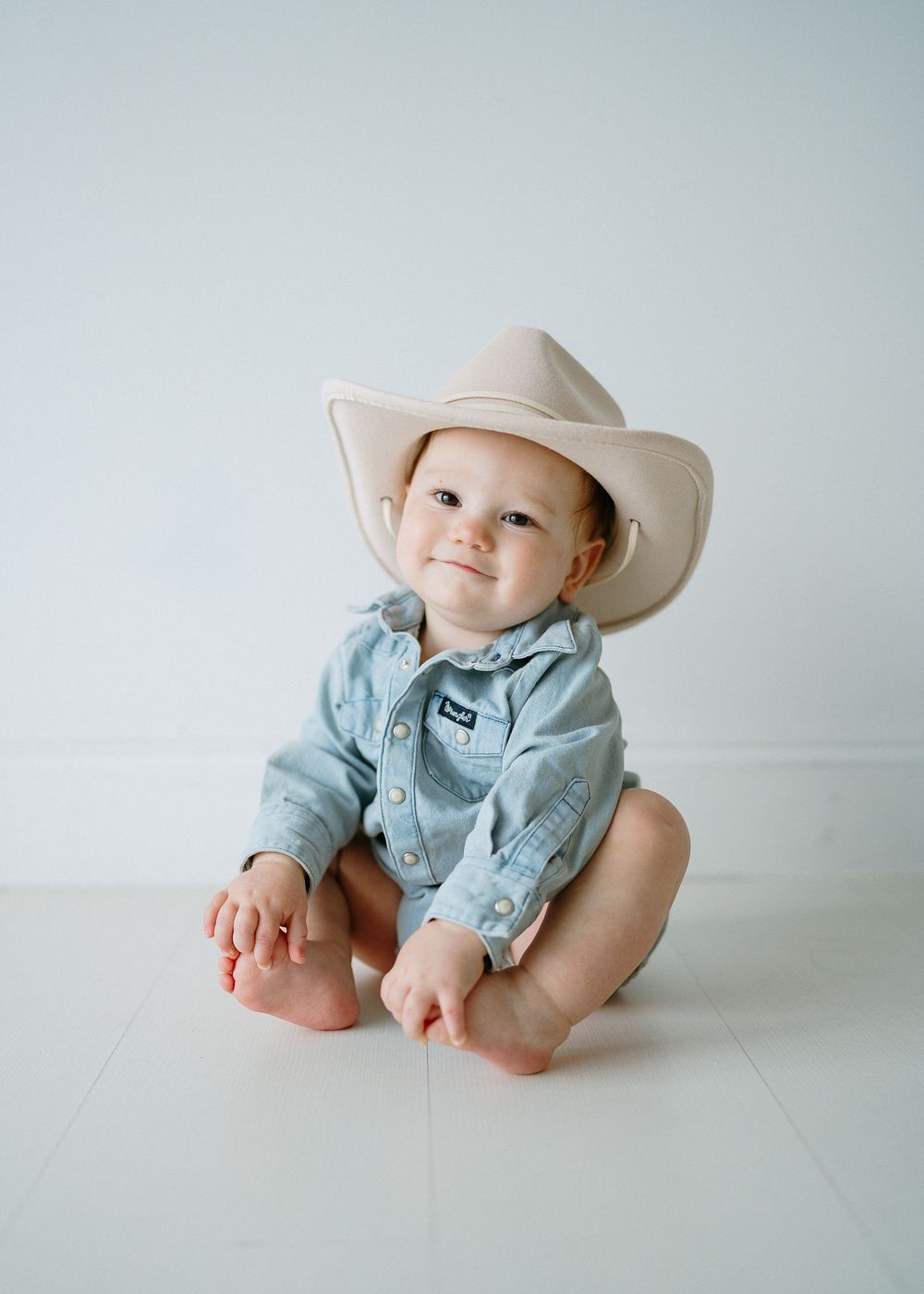Adorable baby wearing a cowboy hat and denim romper sitting on a white floor smiling.