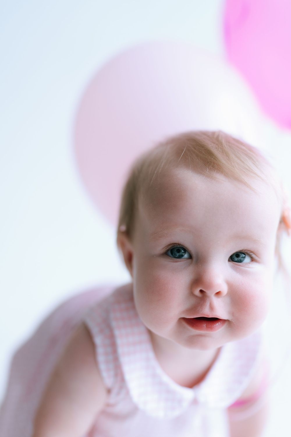 Close-up portrait of blue-eyed baby girl in pink outfit with pink balloons in background.