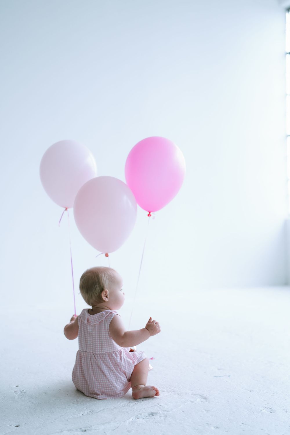 Baby girl in pink romper sitting on white floor holding three pink balloons from behind.