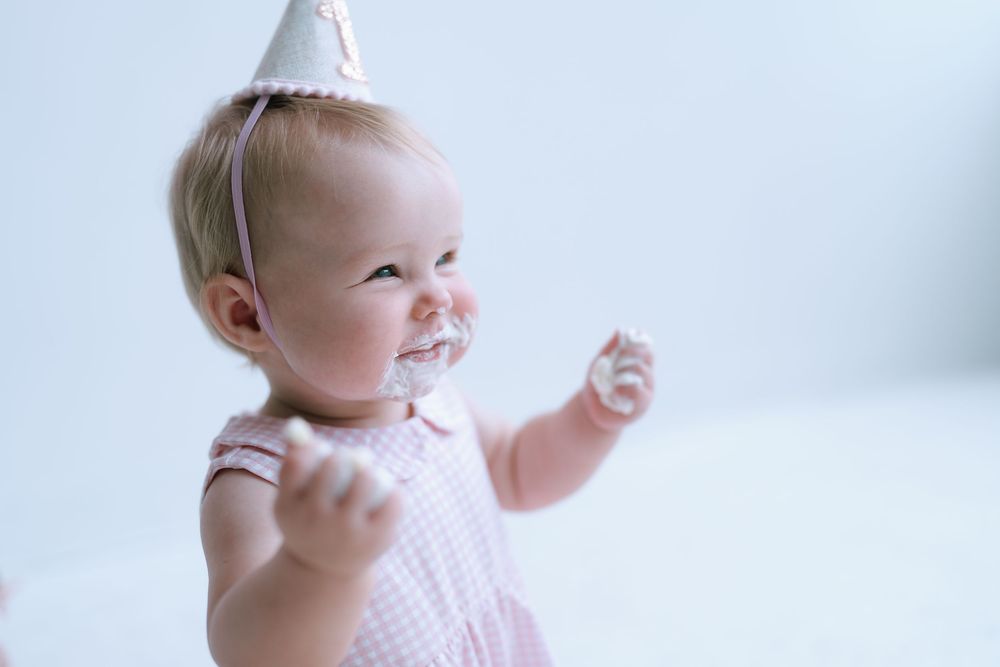 Happy baby girl in pink outfit and party hat with cake smash frosting on her face.