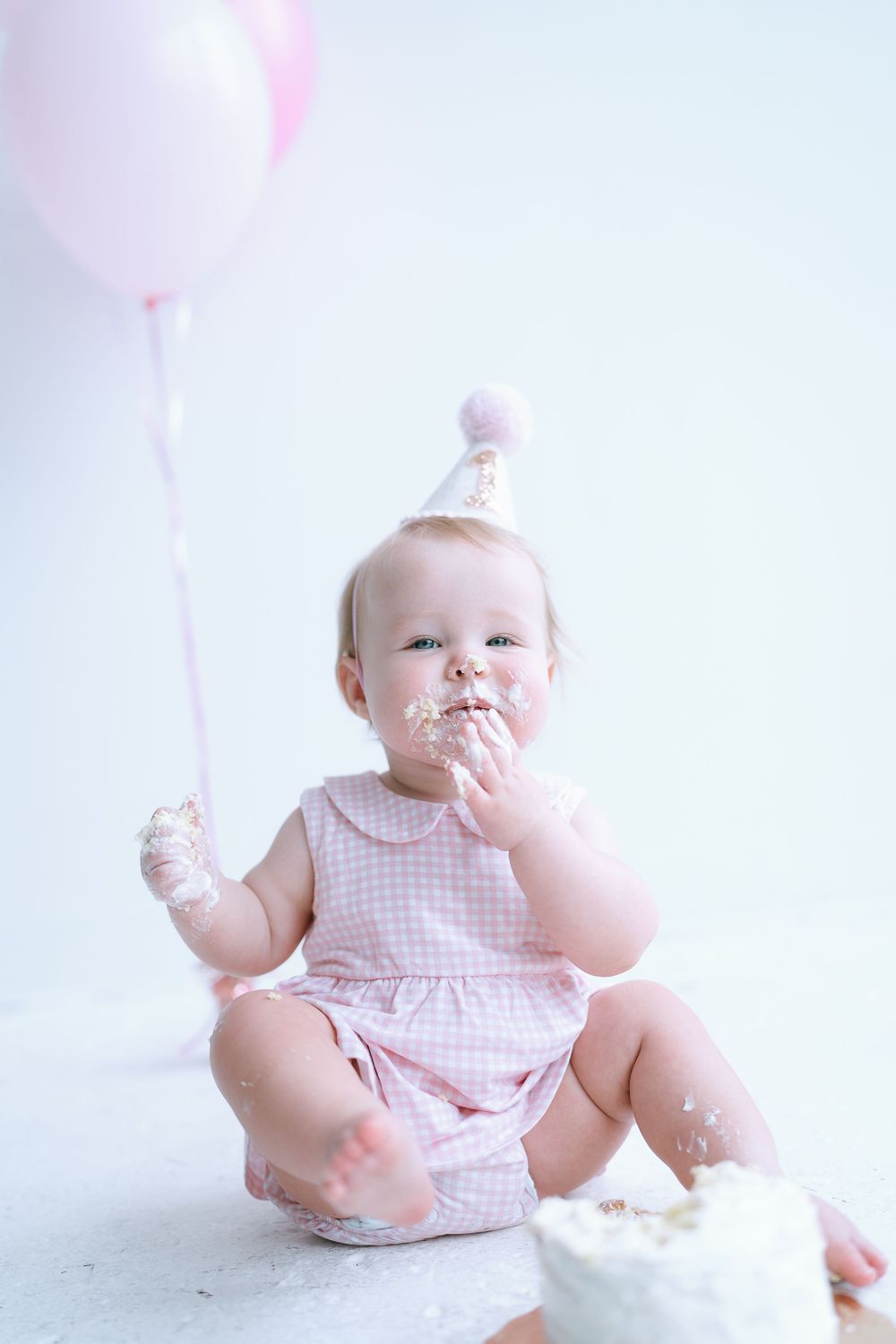 Baby girl in party hat joyfully eating smash cake with frosting on face and pink balloon.