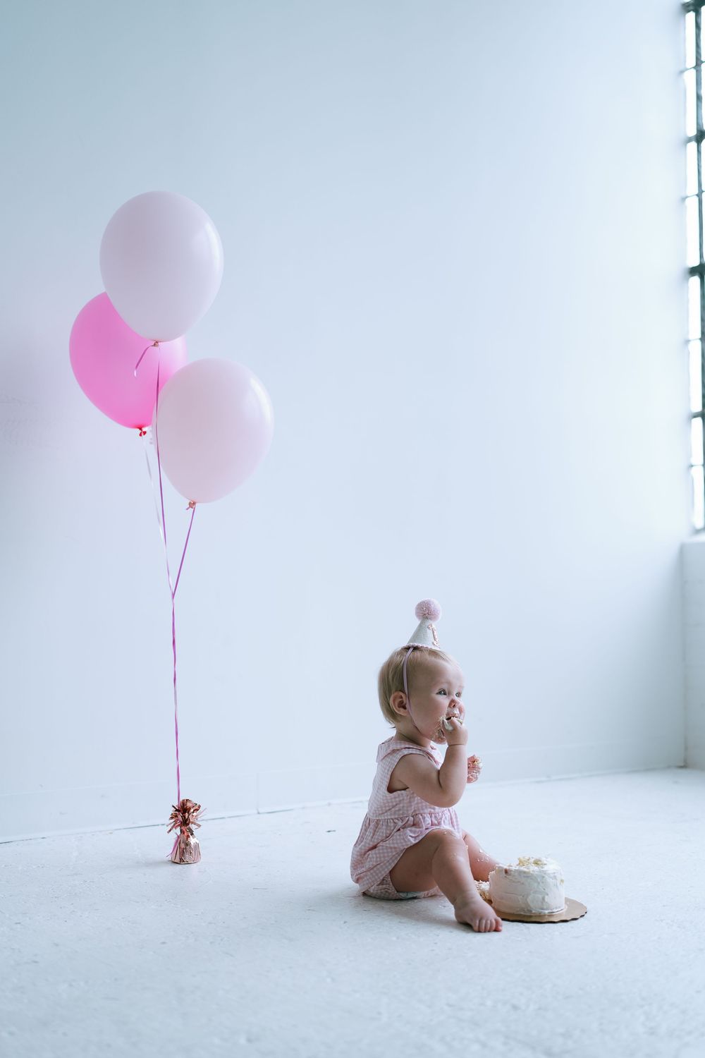Baby girl in pink outfit and party hat sitting beside smash cake with pink balloons nearby.