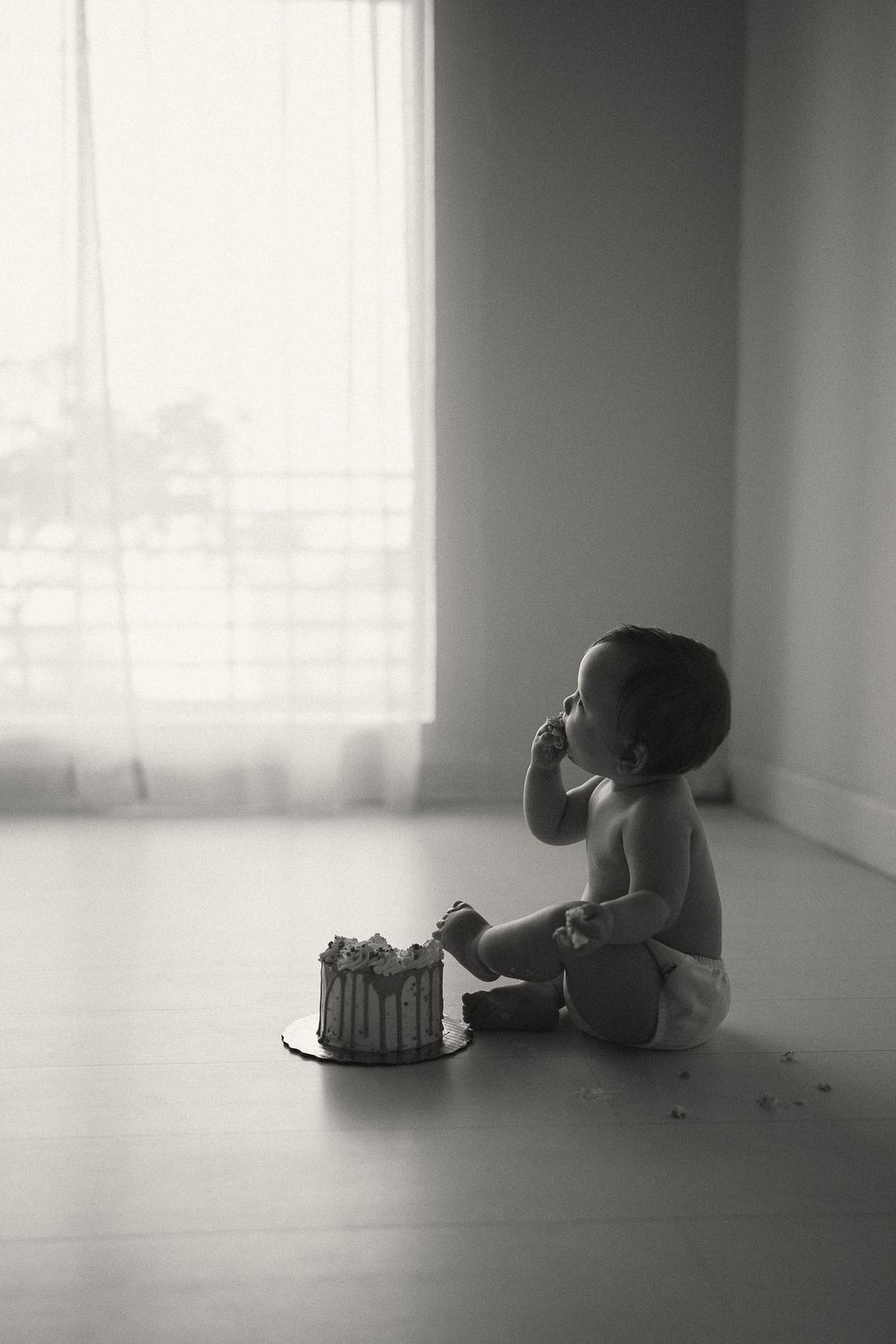 Black and white photo of a baby sitting on the floor eating birthday cake smash near a window.