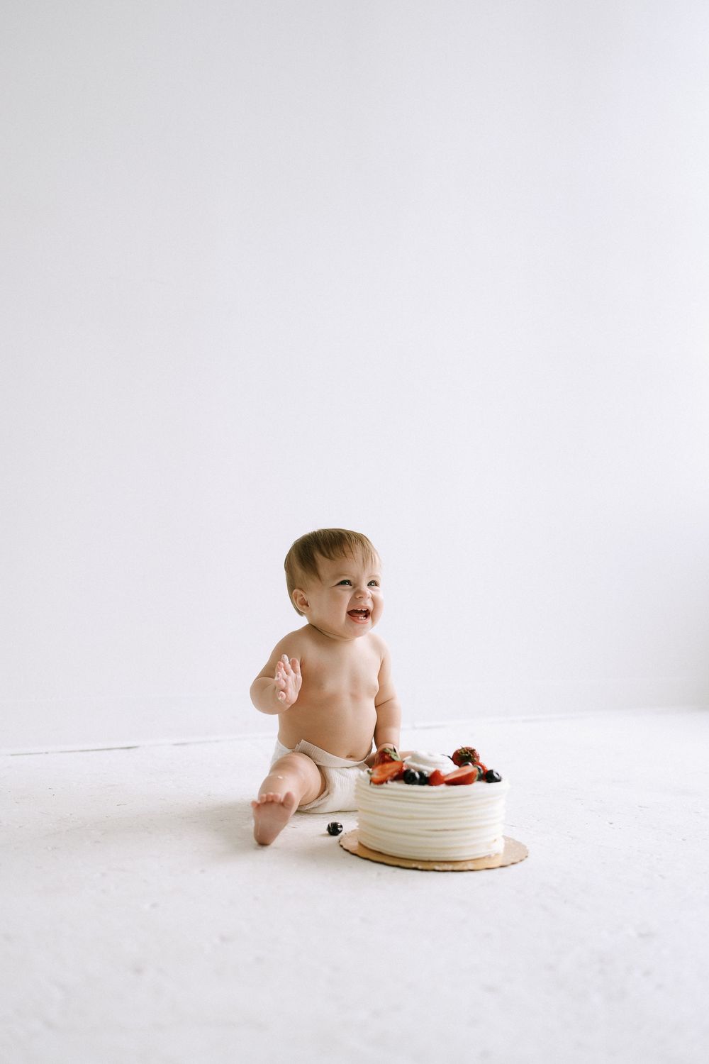 Smiling baby in diaper sitting next to a white frosted cake topped with fresh berries on a white background.