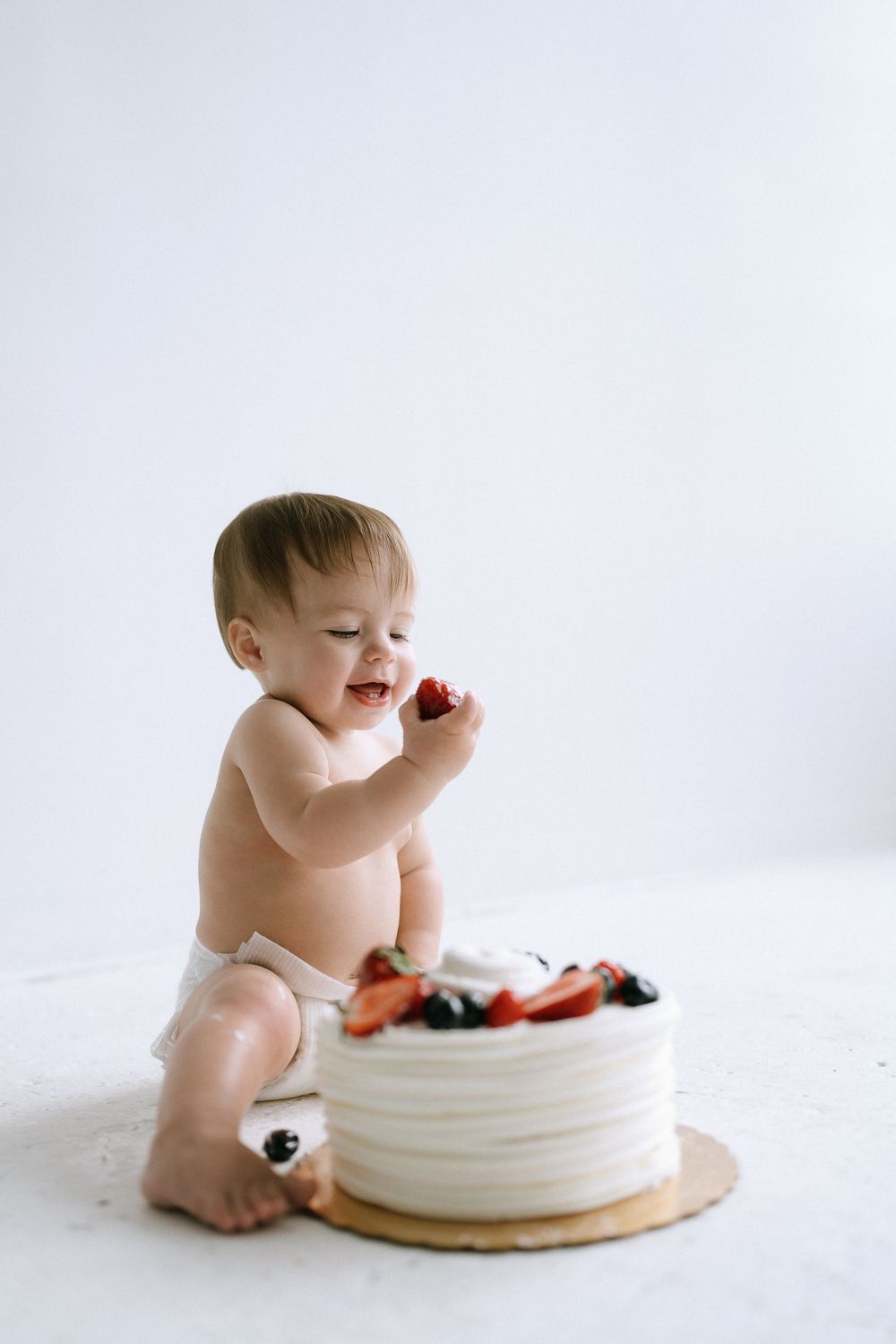 Happy baby in diaper eating strawberries from a white frosted cake decorated with fresh berries.