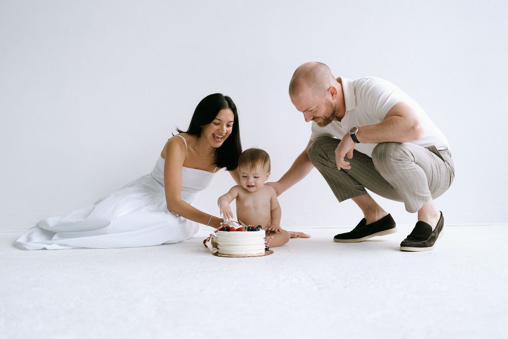 Parents smiling and watching their baby explore a small white birthday cake on a white studio floor.