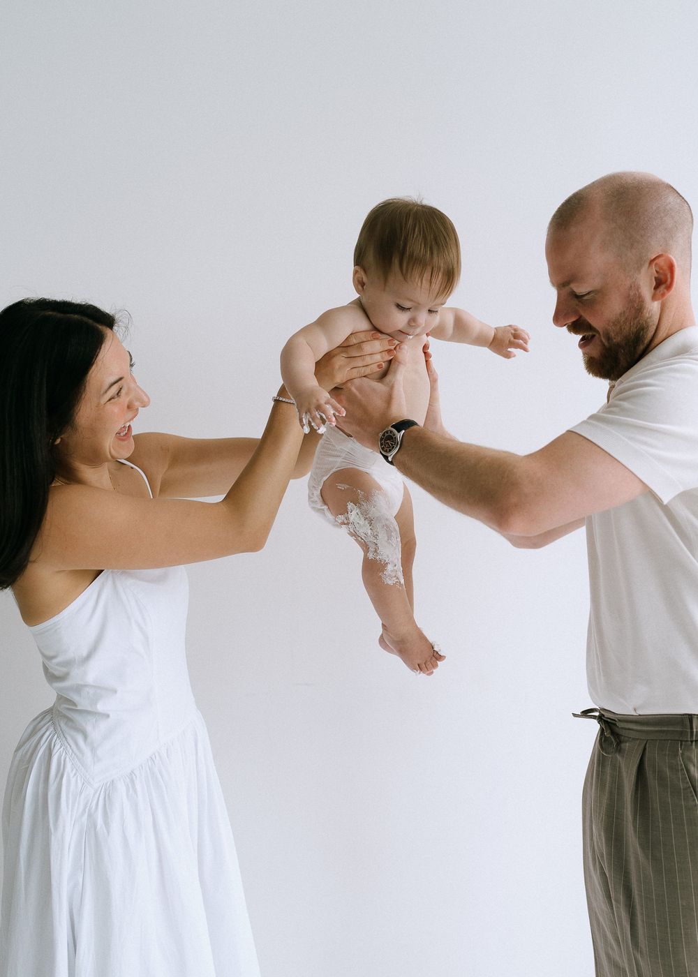 Smiling parents lifting their happy baby in the air against a clean white studio background.