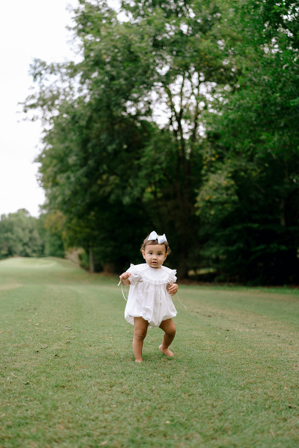 Adorable baby girl in white dress with bow walking barefoot on lush green grass in a park.