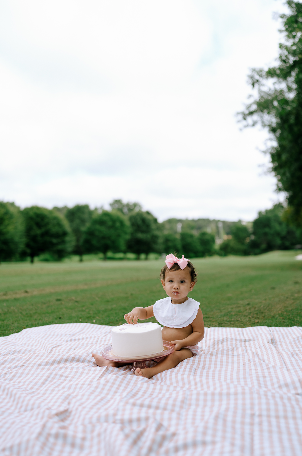 Baby girl in white outfit with pink bow sitting on a blanket with a white cake outdoors.