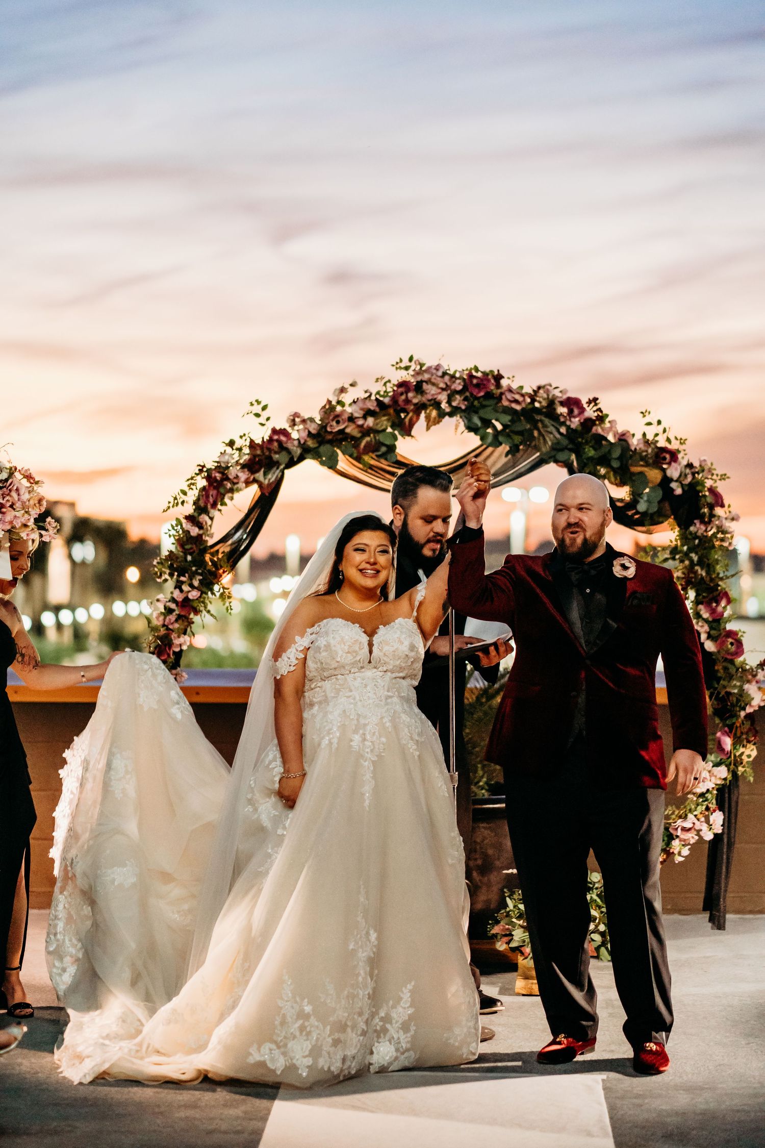 A wedding ceremony under a floral arch at sunset with people celebrating joyfully beneath string lights.