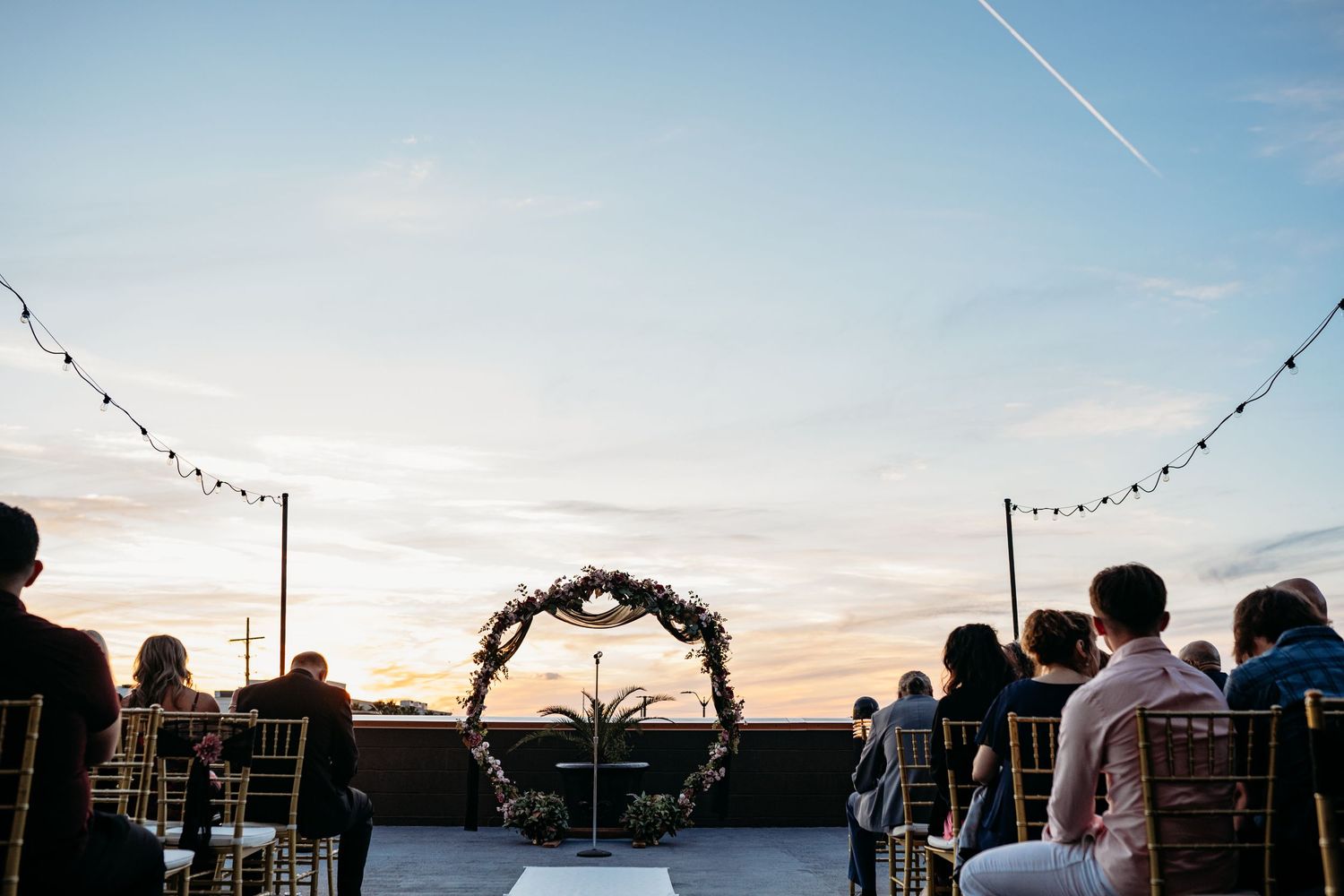 An outdoor rooftop wedding ceremony setup at sunset with string lights and decorative arch against a vibrant sky.