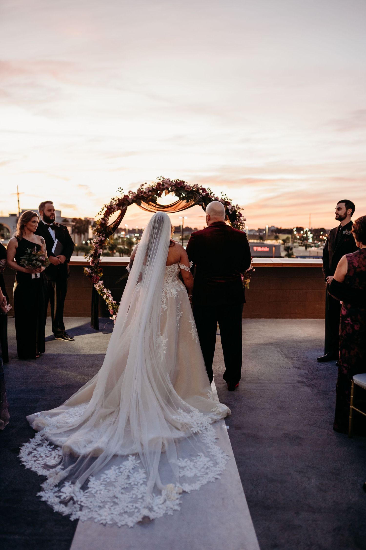 A wedding ceremony taking place on a rooftop at sunset with a floral arch and flowing lace veil in view.