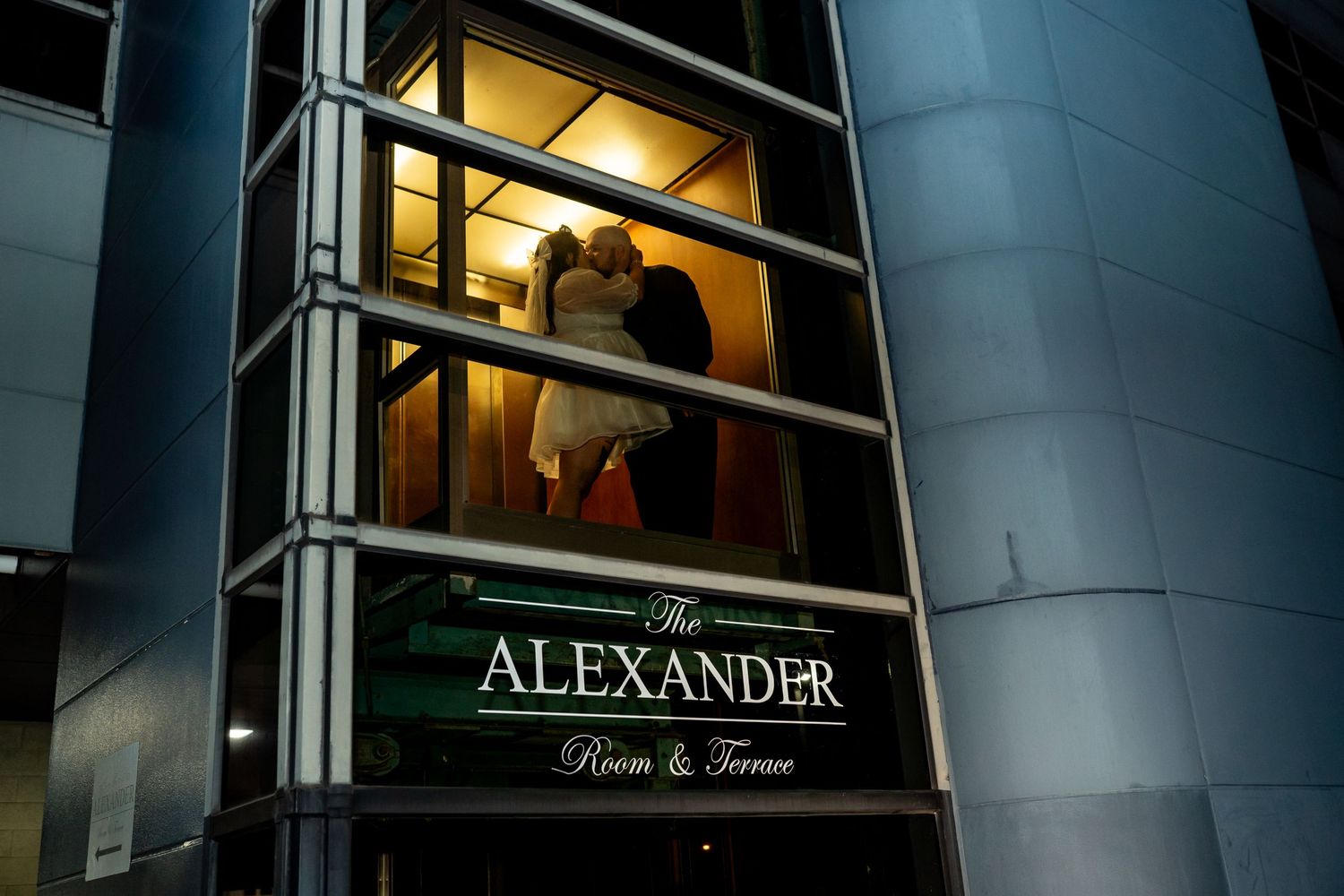 An illuminated architectural window display shows the Alexander sign with warm lighting at night.