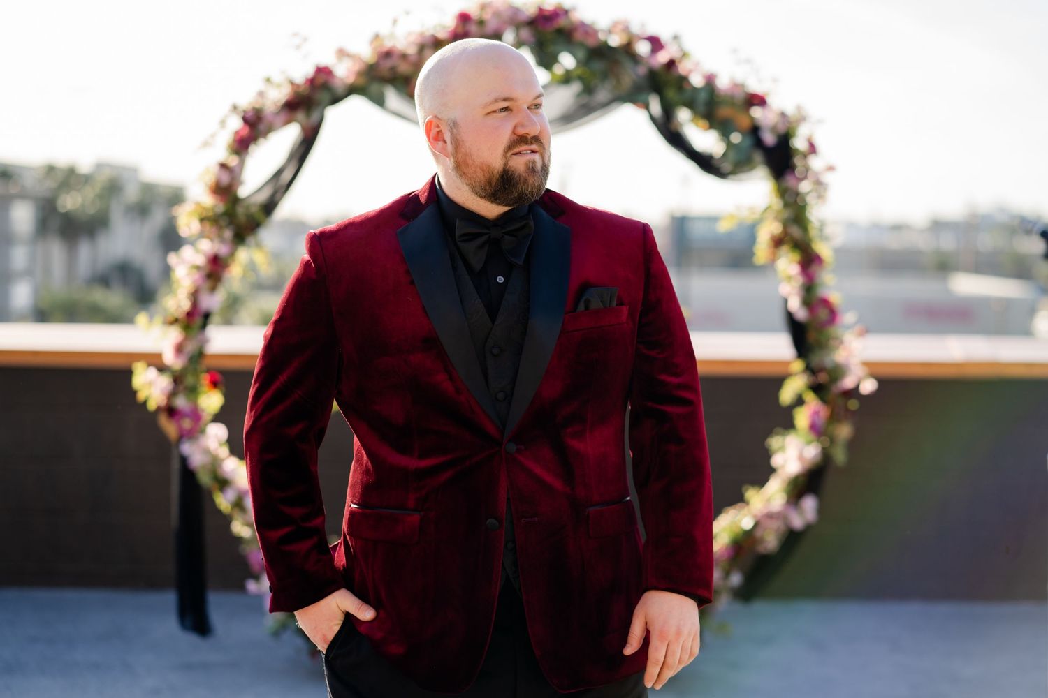 A person in a burgundy velvet tuxedo jacket stands in front of a floral wedding arch on a rooftop.