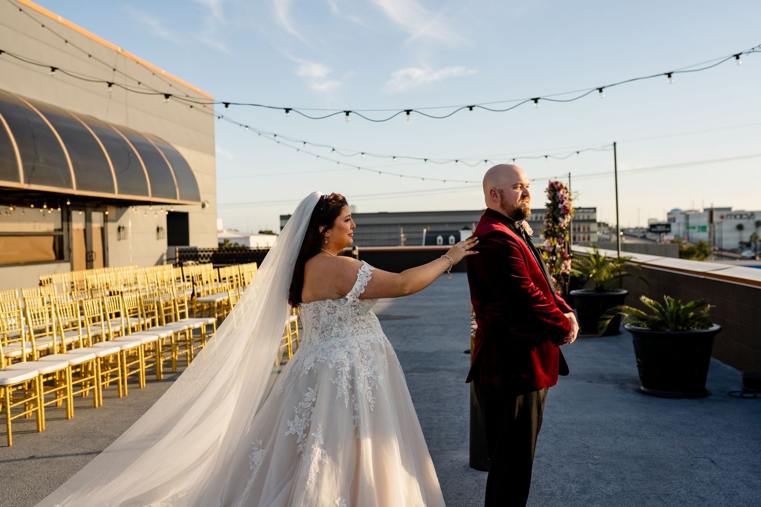 A couple shares their first look moment at their rooftop wedding venue with string lights and golden chairs.