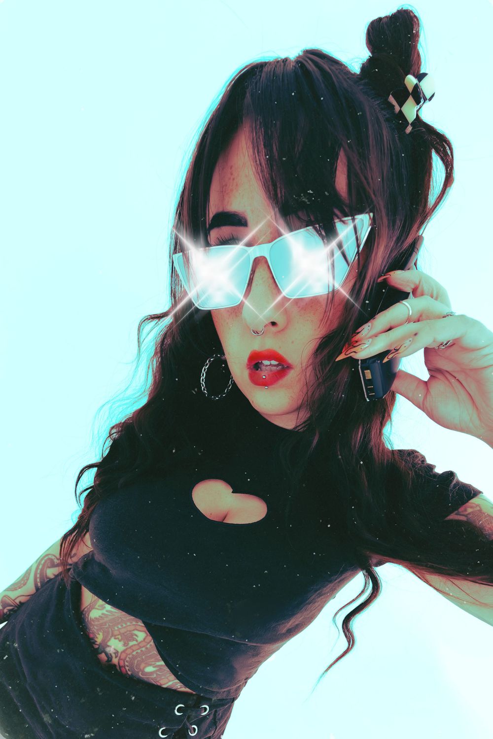 Woman in black crop top with heart cutout, sparkly oversized sunglasses, hoop earrings, and tattoos poses against light blue background.