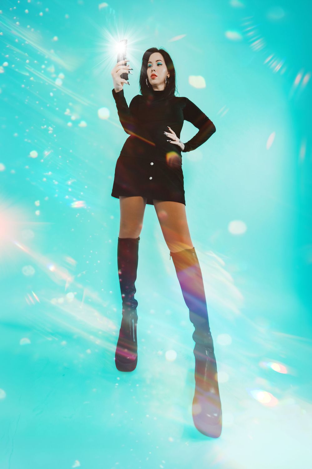 Woman in black mini dress and rainbow thigh-high boots takes selfie against sparkling turquoise background.