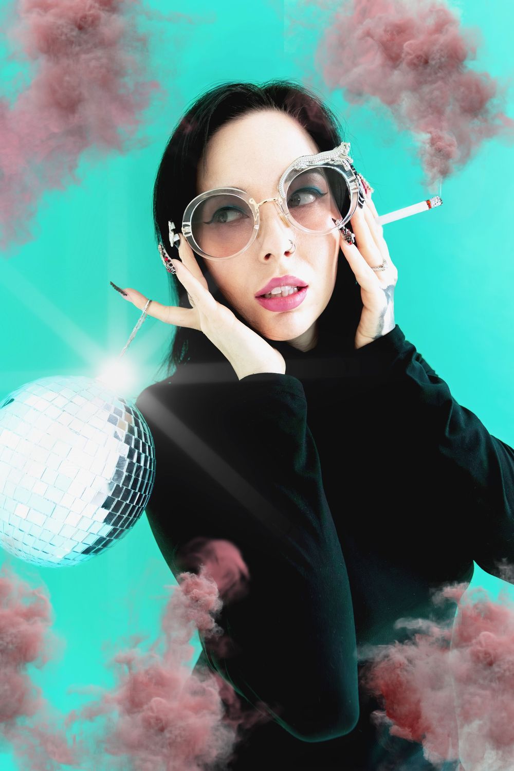 Stylish woman in round sunglasses holds cigarette near disco ball with pink smoke clouds on teal background.