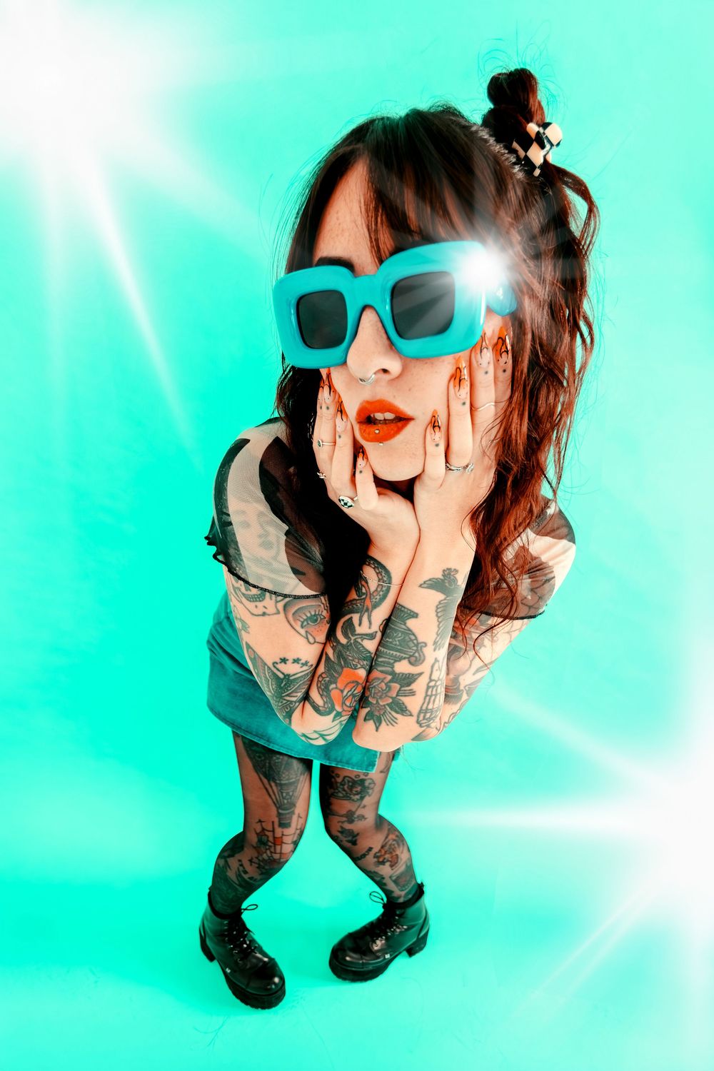 Tattooed woman in teal oversized sunglasses and mini dress poses with hands on cheeks against bright turquoise background.
