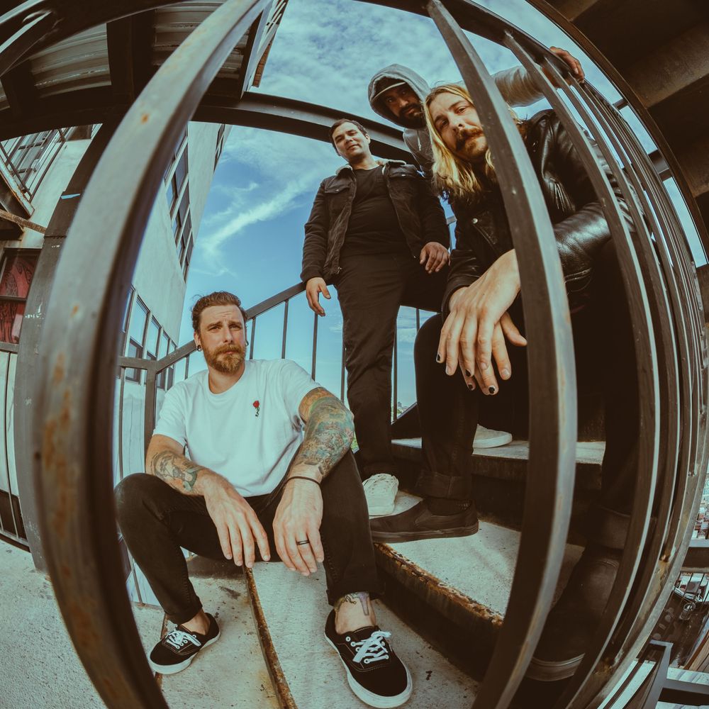 Three tattooed band members pose on outdoor stairs using a fisheye lens under a bright blue sky.