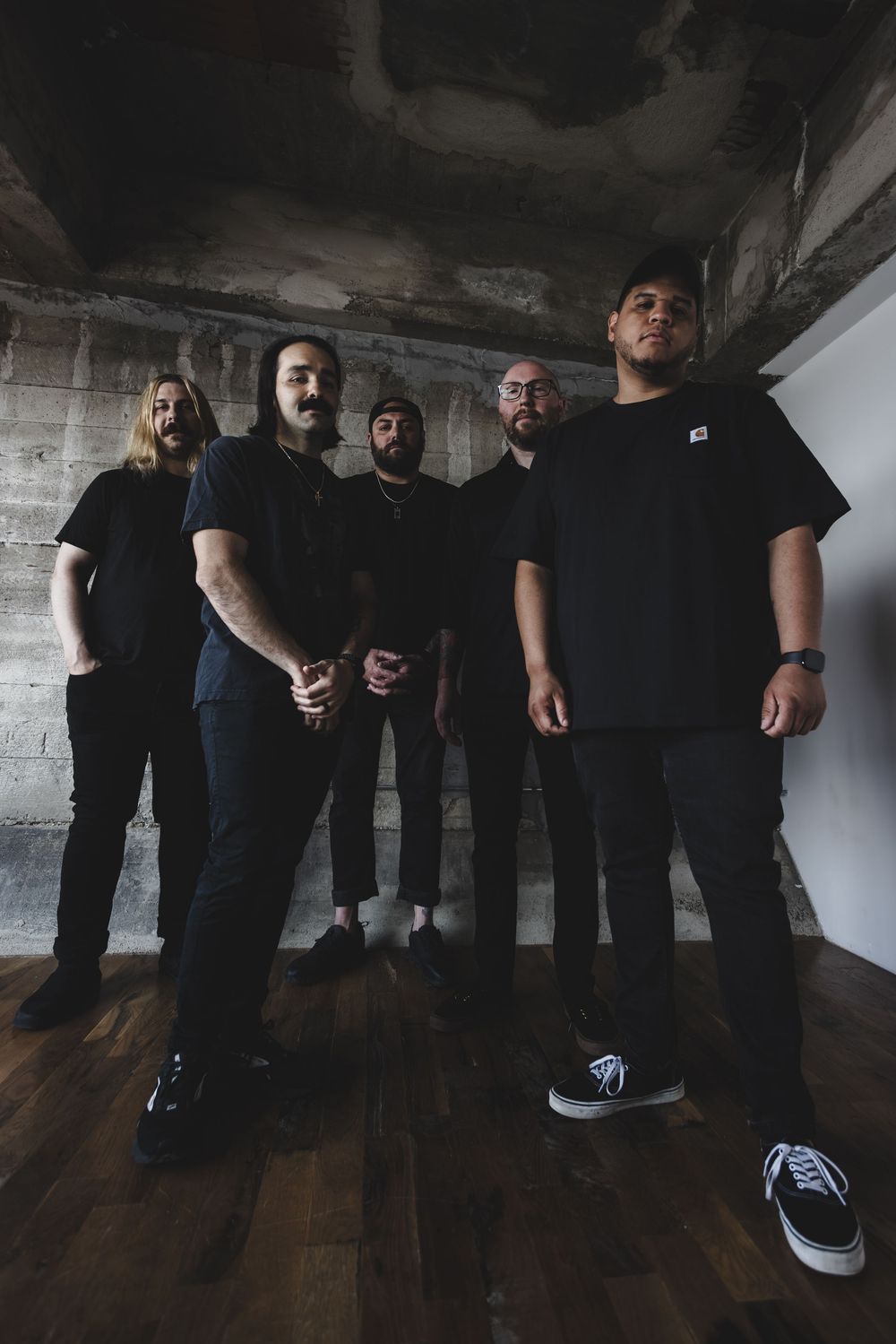 Five musicians in black clothing pose together in a moody concrete room with wooden floors.