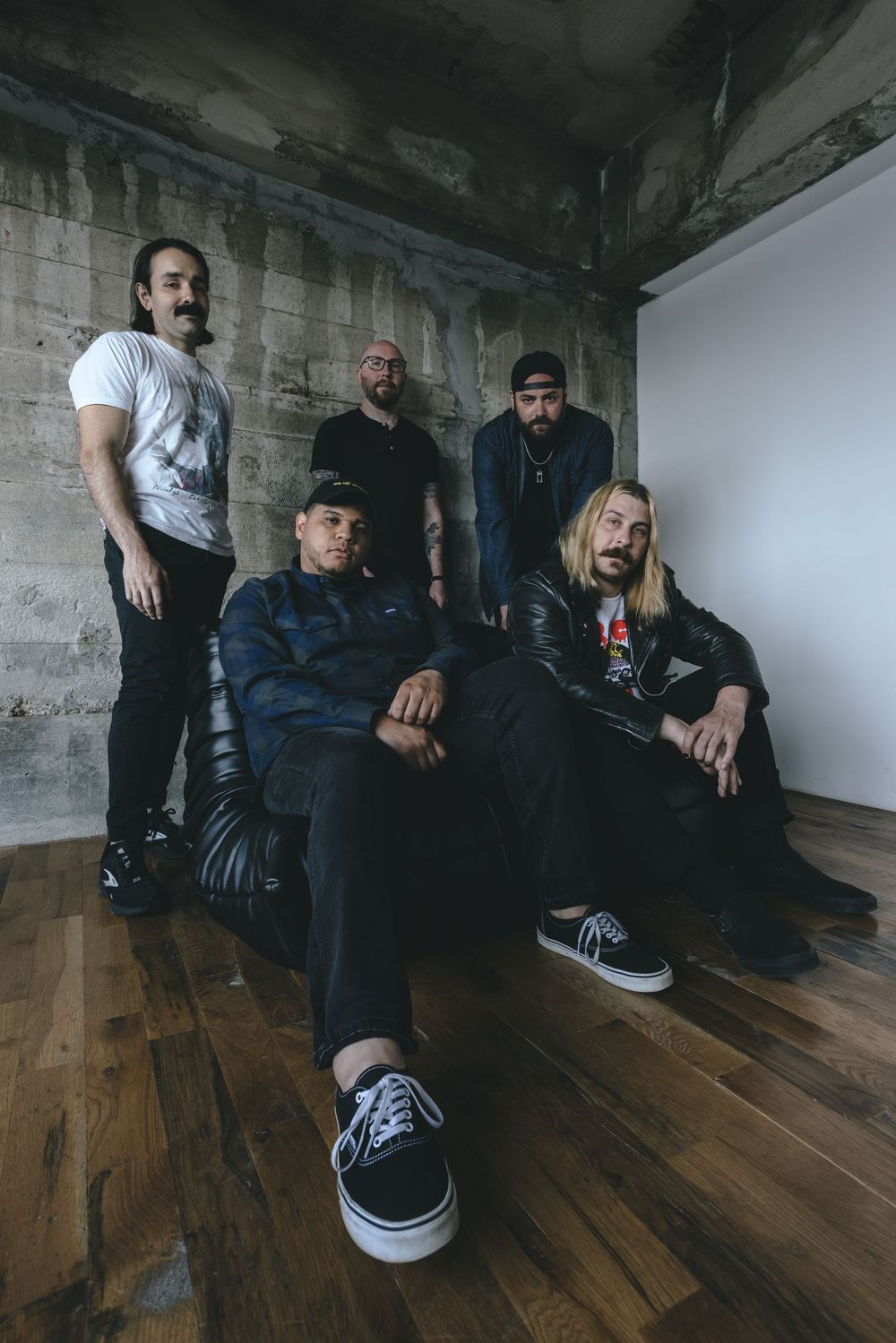 Five members of a rock band pose in a rustic concrete room with wooden floors, some seated and some standing.