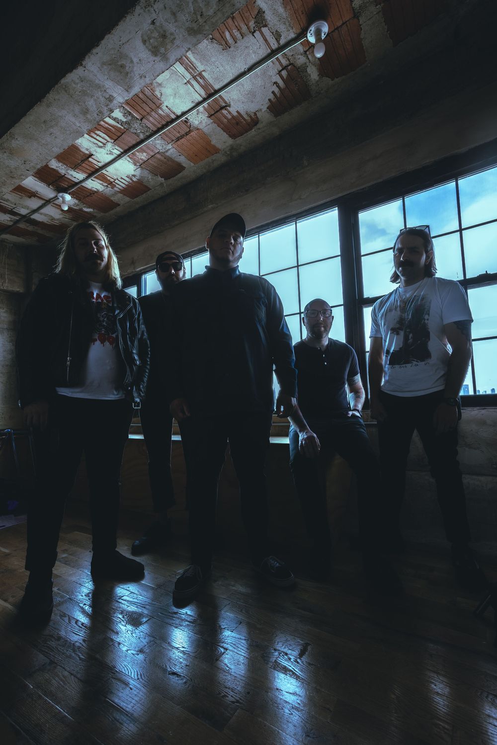 Silhouettes of five band members standing in a dimly lit industrial room with large windows and wooden floors.