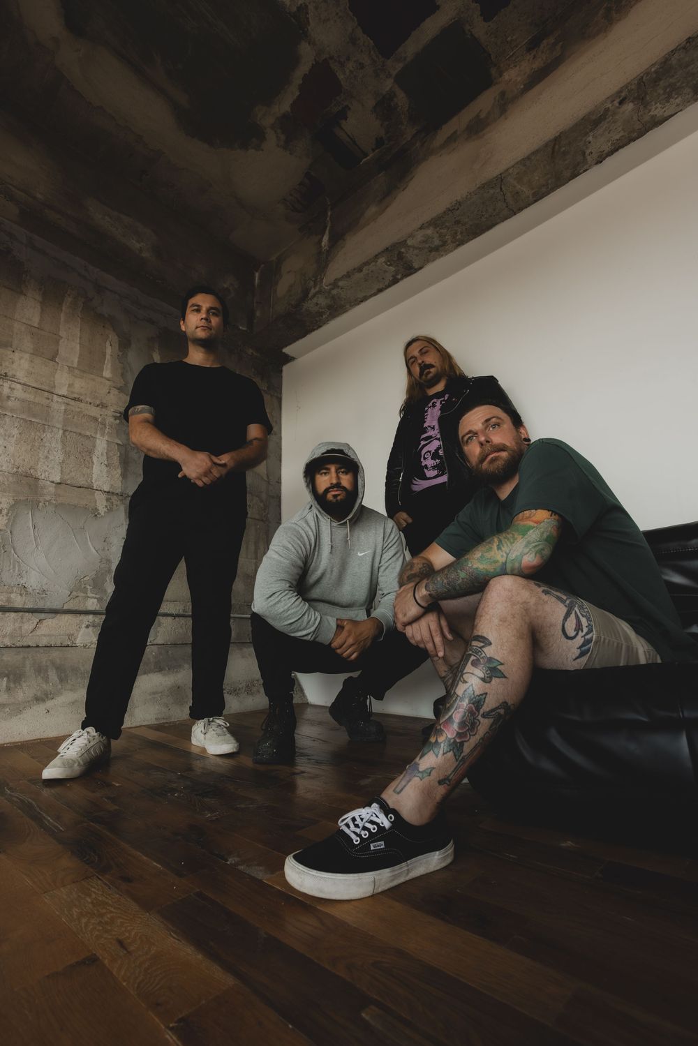 Four tattooed band members pose in a rustic industrial space with concrete walls and hardwood floors.
