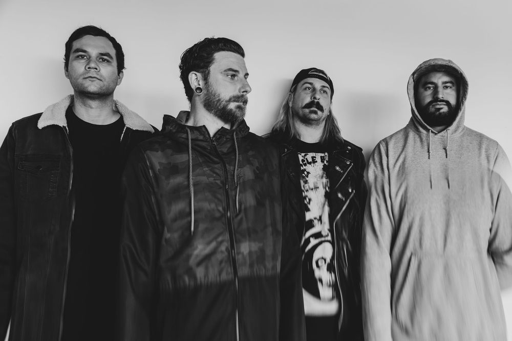 Four men pose together in a black and white band photo against a light background, wearing casual and edgy streetwear.