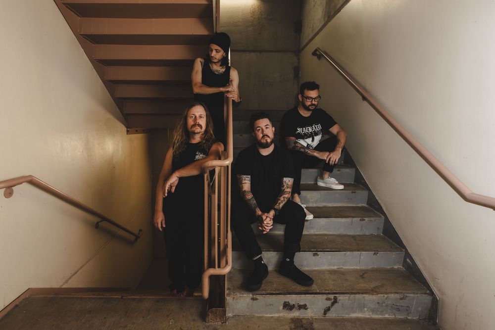 Three band members pose on concrete stairs in a dimly lit stairwell, all wearing black clothing.