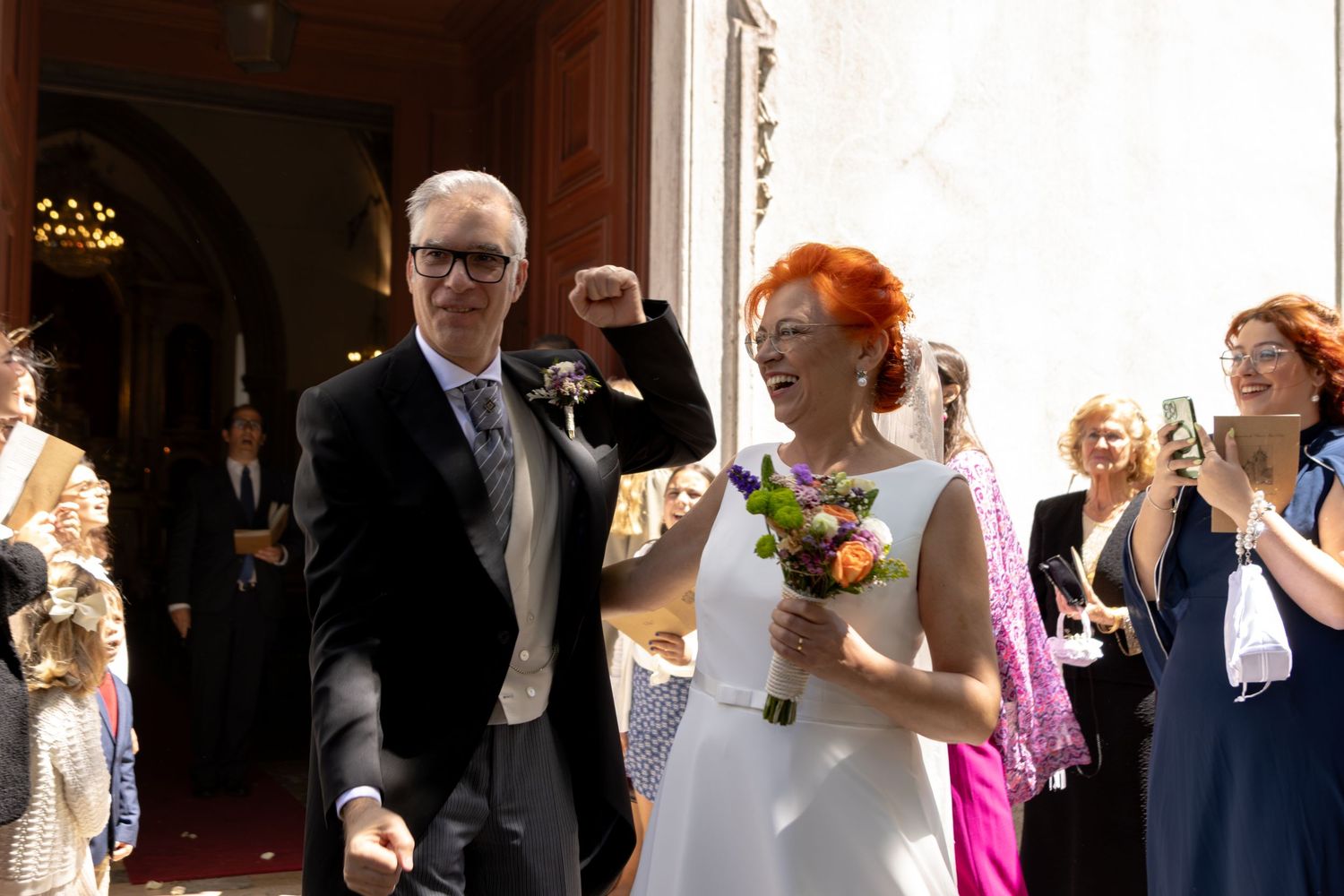 Newlyweds exit church ceremony while guests celebrate and throw confetti in sunlight.