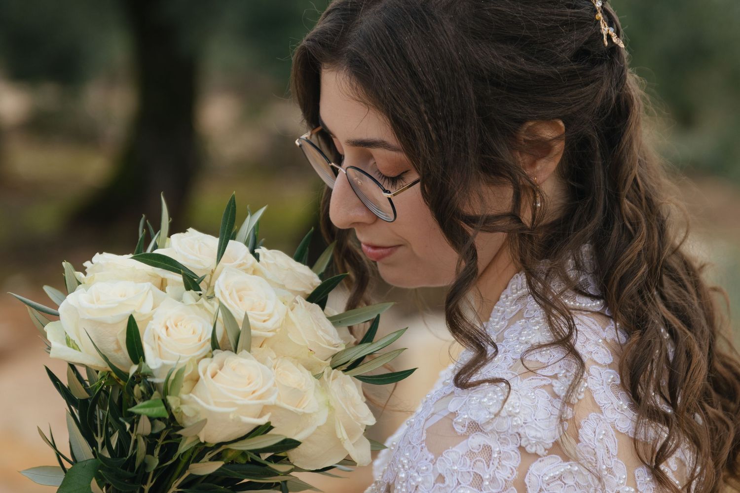 A bride in a white lace dress with long sleeves holds a bouquet of white flowers against a natural outdoor backdrop.