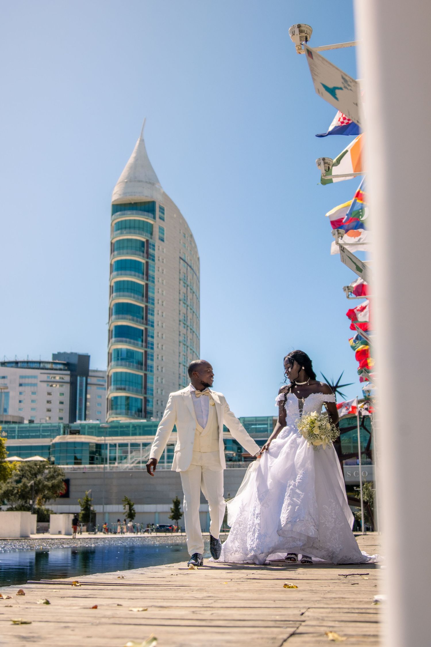 Wedding couple poses on modern architecture plaza with glass buildings and colorful flags.