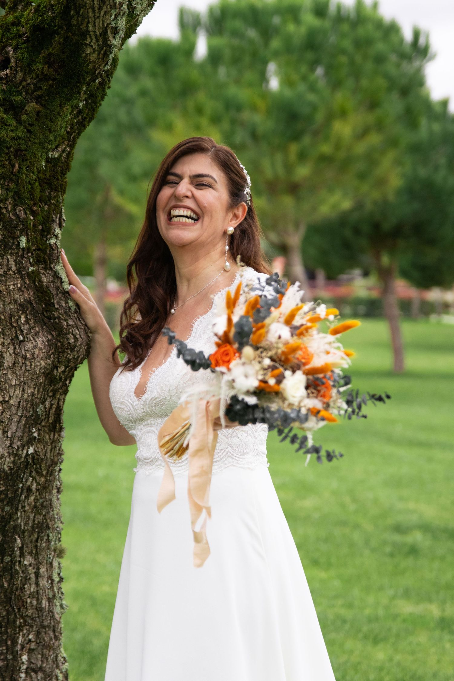 A bride in a white lace dress laughs joyfully while holding an orange and white bouquet in a garden setting.