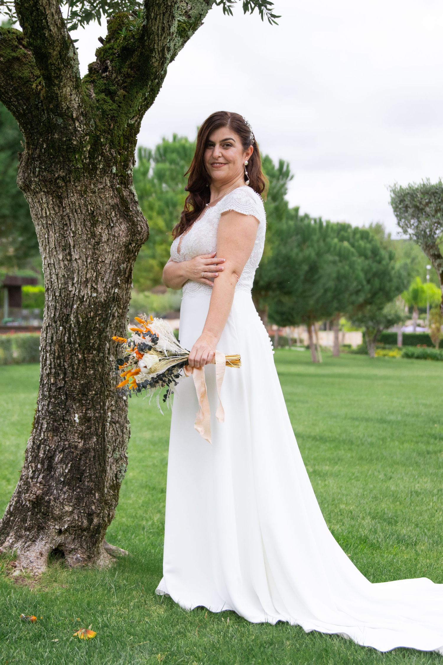 A bride poses with her bouquet against a tree trunk in a lush green park setting.